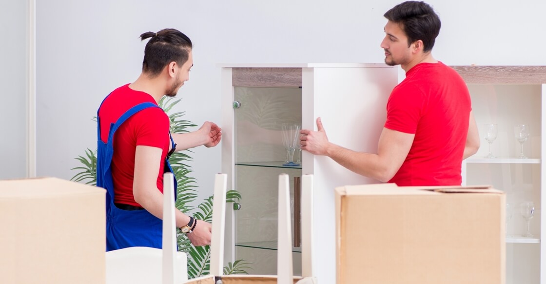 Two moving men carefully handling a glass cabinet, preparing it for transport, with packing boxes in the background.