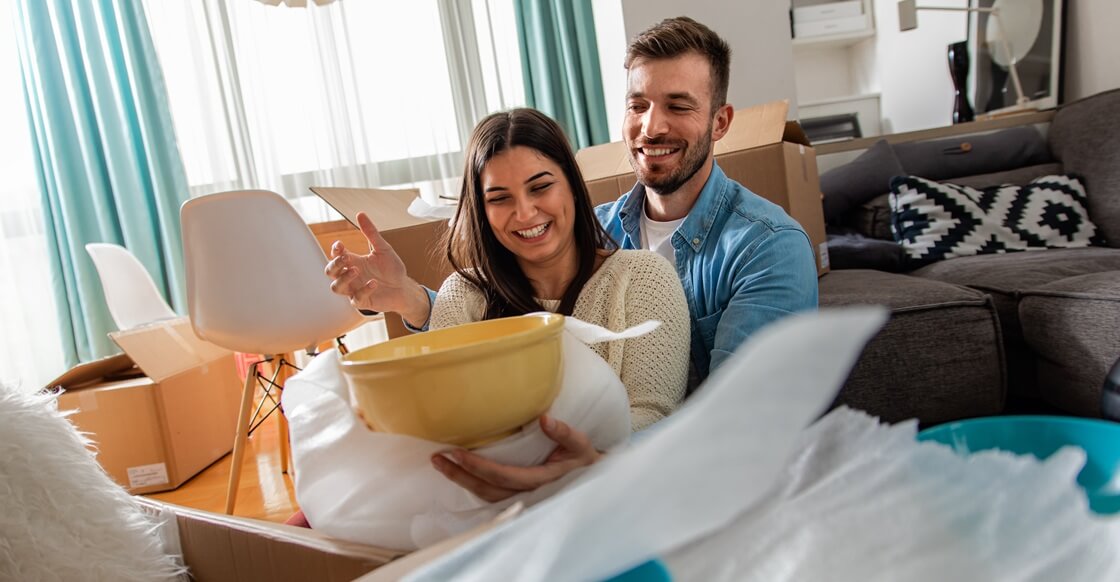 Smiling young couple unpacking boxes in their new home, holding a wrapped yellow bowl while sitting on the floor surrounded by moving supplies.